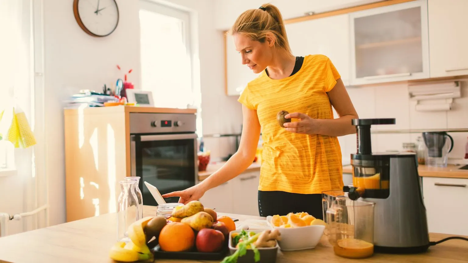 Woman using a juicer in her kitchen while looking at a recipe on her laptop