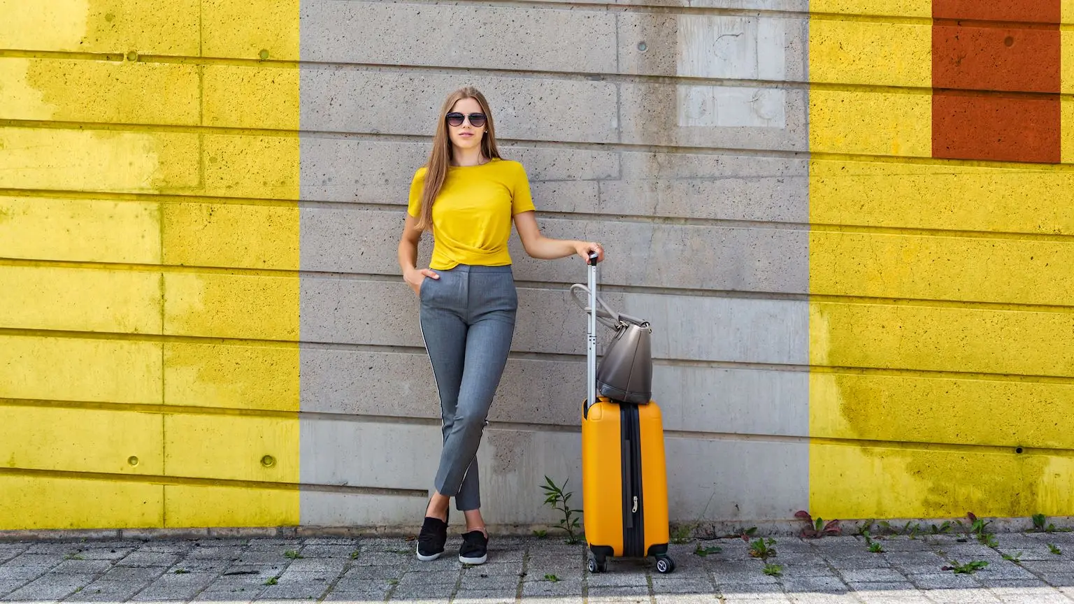 Woman wearing a yellow t-shirt standing with a yellow suitcase in front of a yellow painted wall