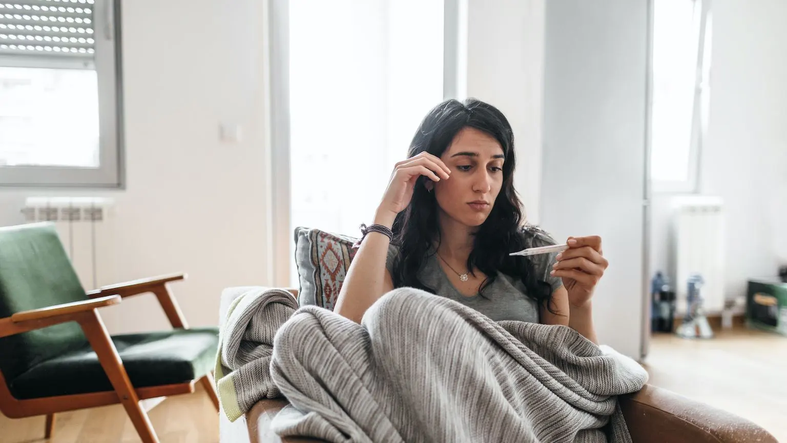 Woman sitting in a chair, wrapped in a blanket, taking her temperature