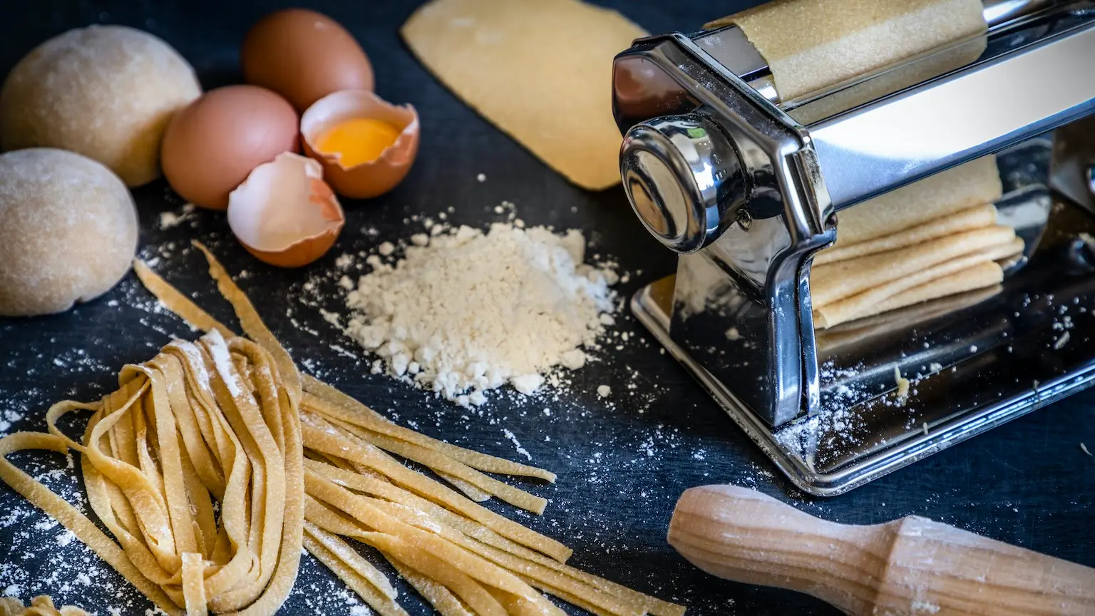 Front view of a homemade pasta machine surrounded by the ingredients for making pasta like eggs and flour. Low key DSLR photo taken with Canon EOS 6D Mark II and Canon EF 24-105 mm f/4L