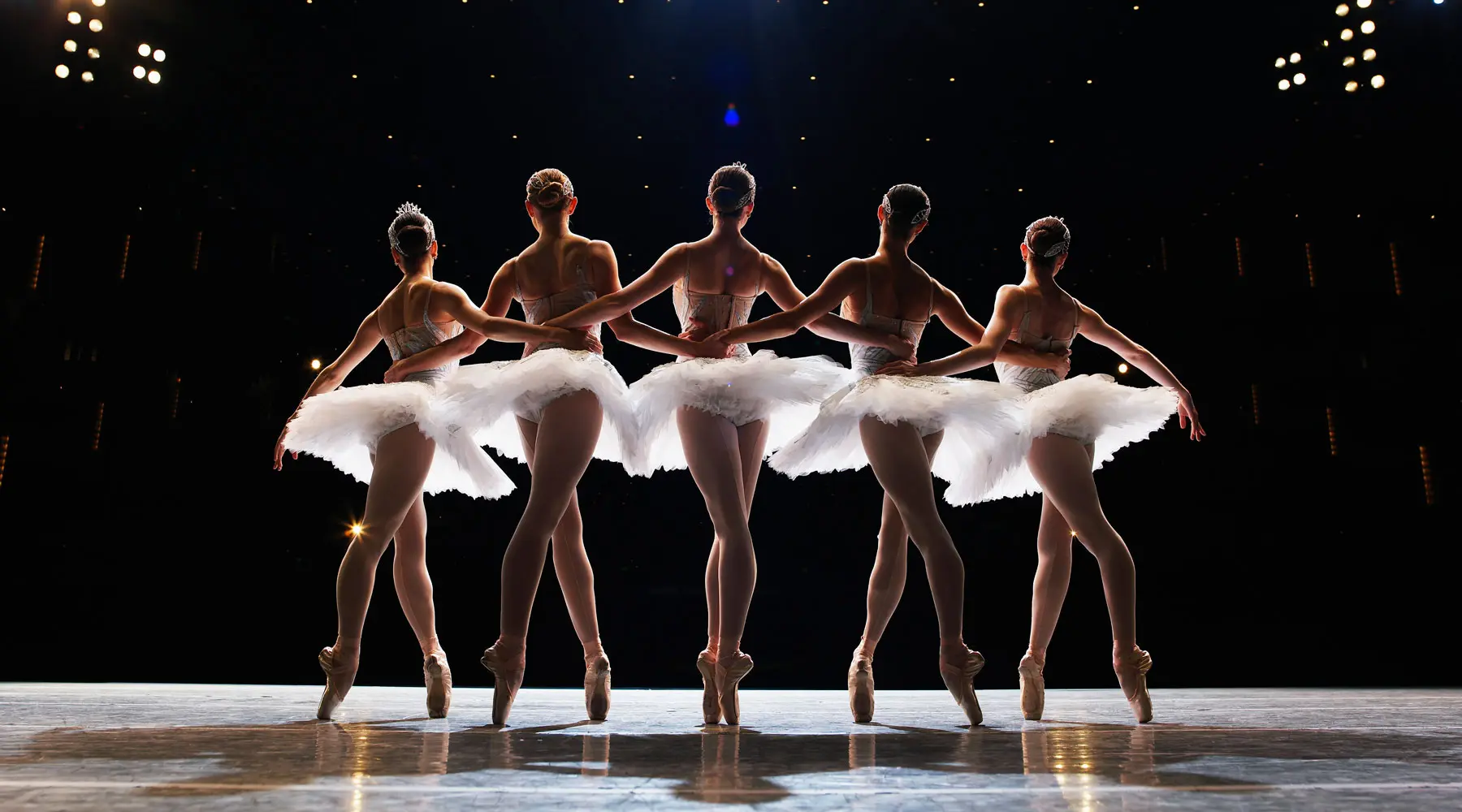 Five ballerinas en pointe on stage, arms around each other, rear view