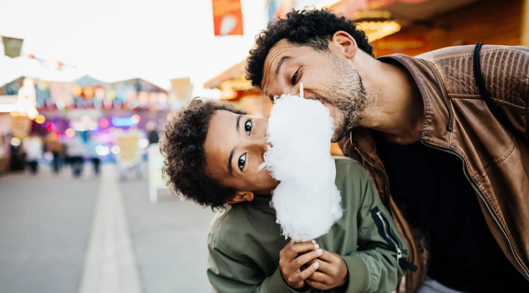 A playful father and son sharing a bloom of candy floss while spending the day at the fun fair together.